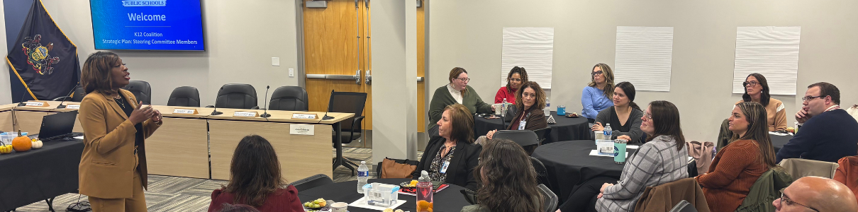 Room full of people sitting around round tables, being led in discussion by CAO ShaQuita Gatewood at front left.